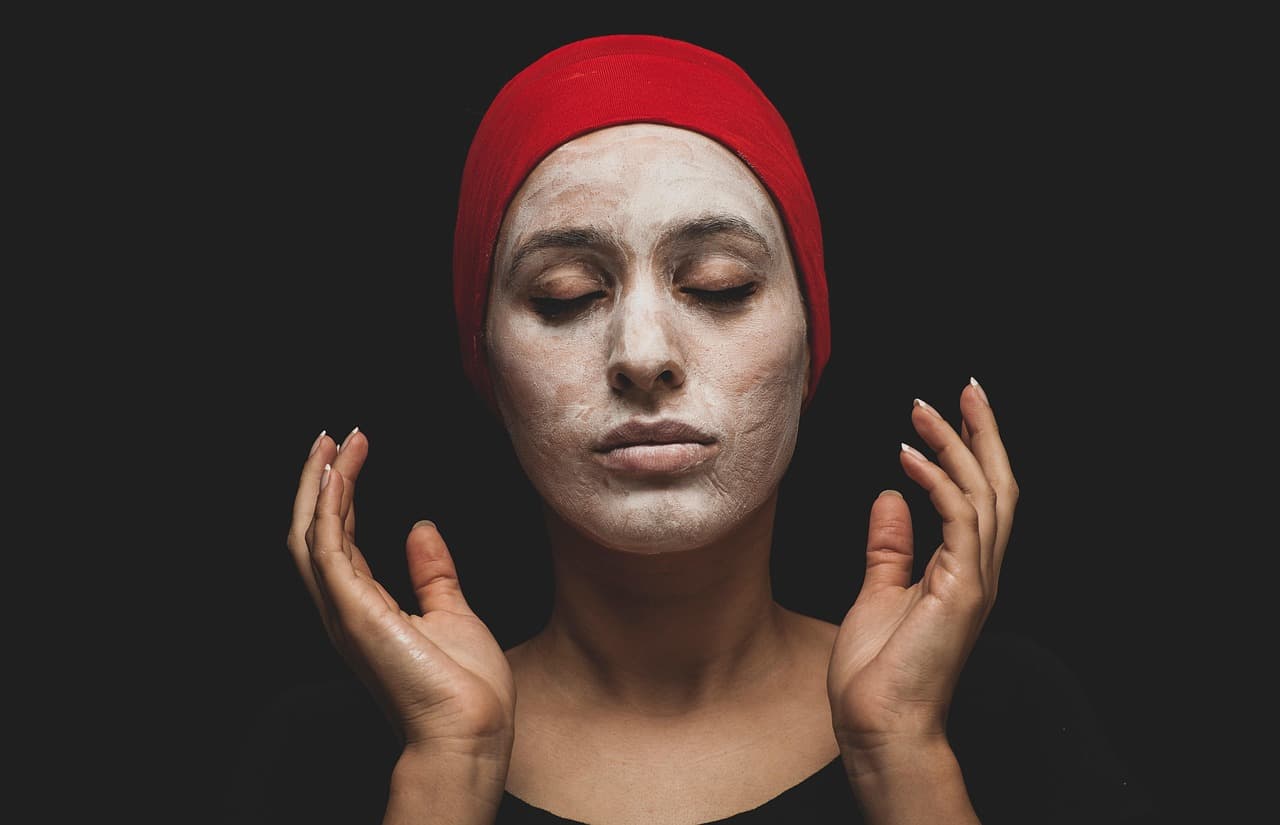 A woman with a clay mask on, eyes closed, hands raised beside her face